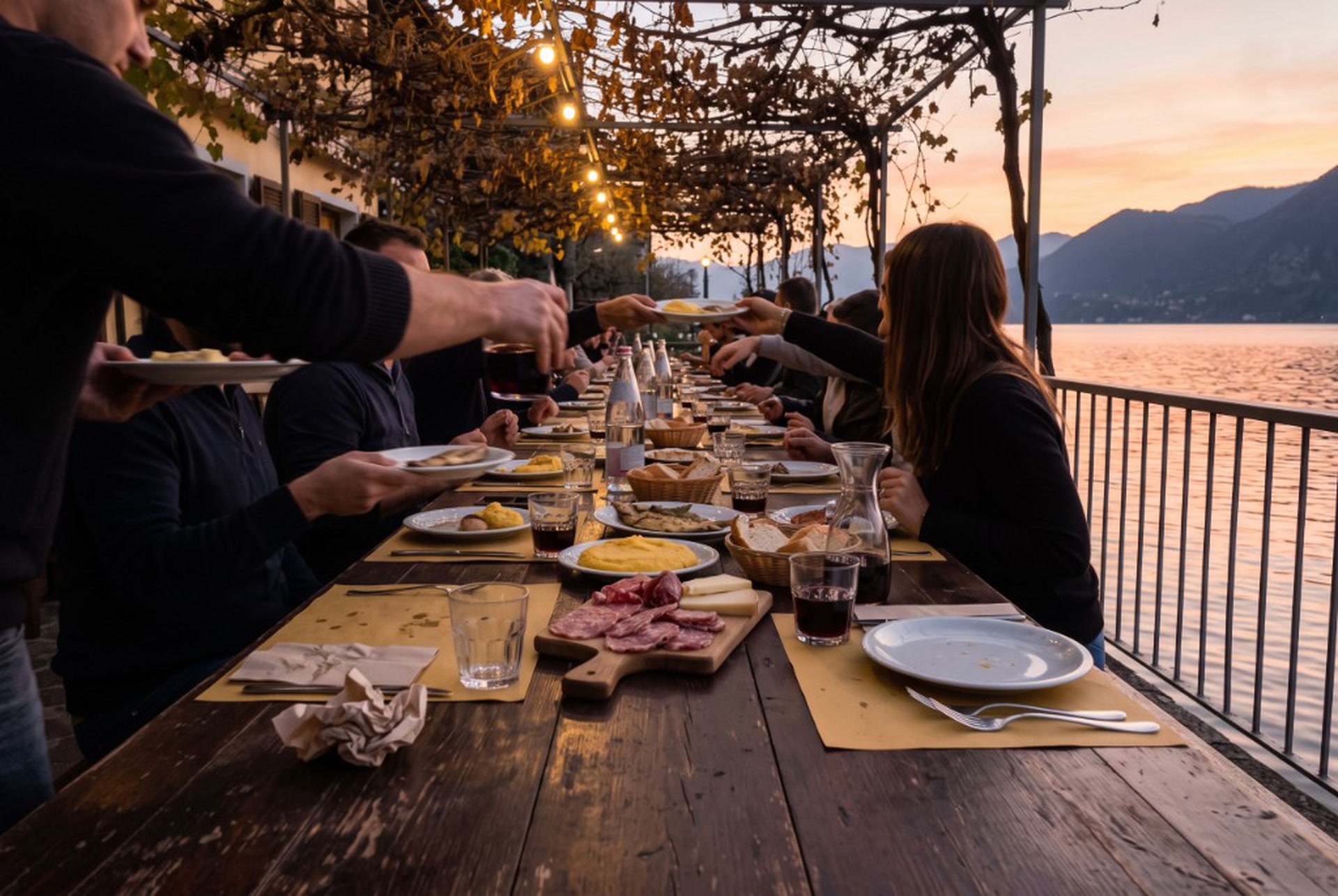 Cena lunga sul lago al tramonto, tavola condivisa sotto le luci